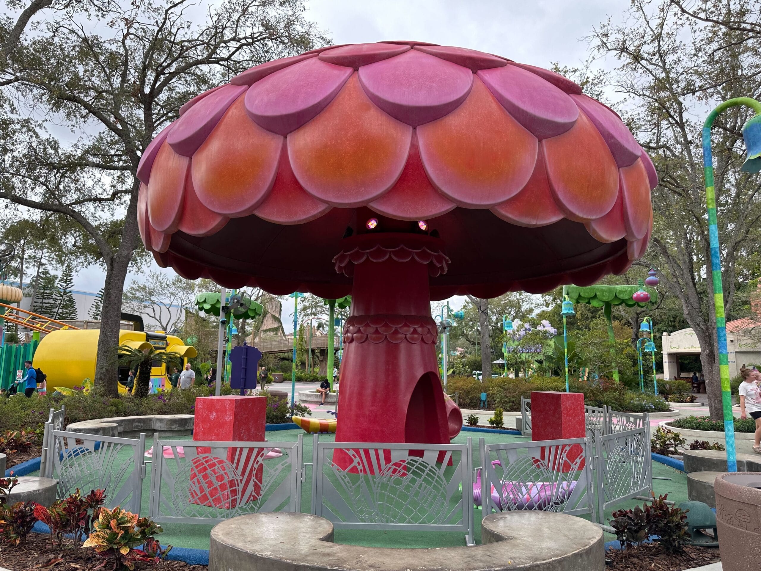 Large red mushroom-shaped playground surrounded by a fence in an outdoor park area with trees and paths.