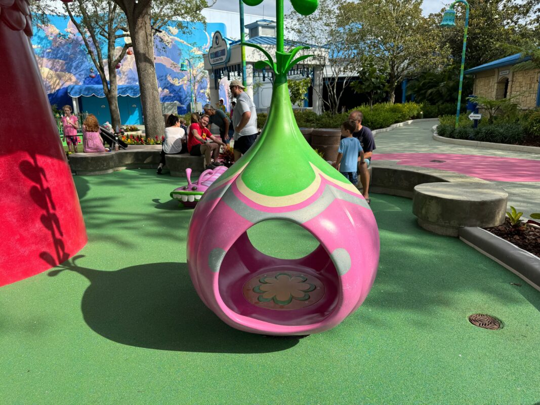 A bright pink and green playground structure shaped like a bulbous pod reminiscent of DreamWorks Land, with children and adults interacting in the background on a sunny day.