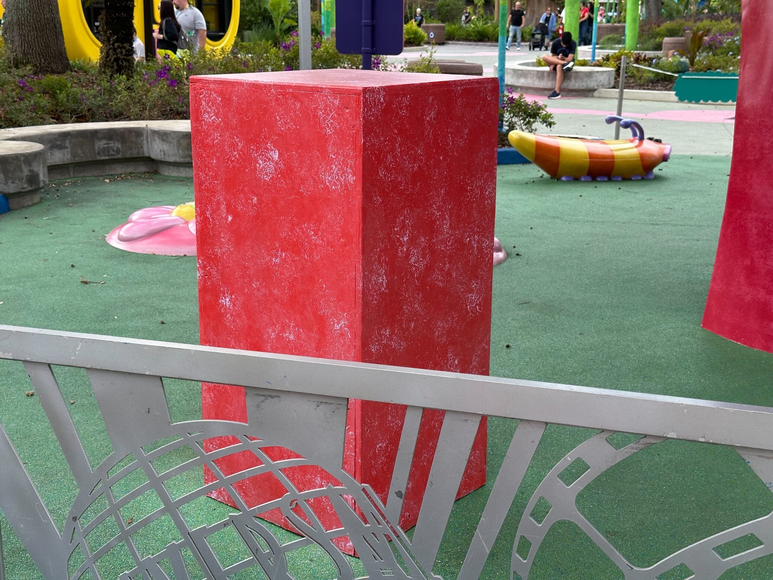 A tall red rectangular block stands on a green playground area surrounded by a decorative fence. In the background you can see a colorful playground structure and people sitting on benches.