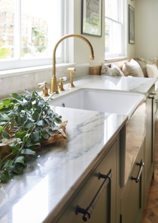 Close-up of a kitchen sink and veined sideboards with gold taps. There is a blurred sofa in the background.