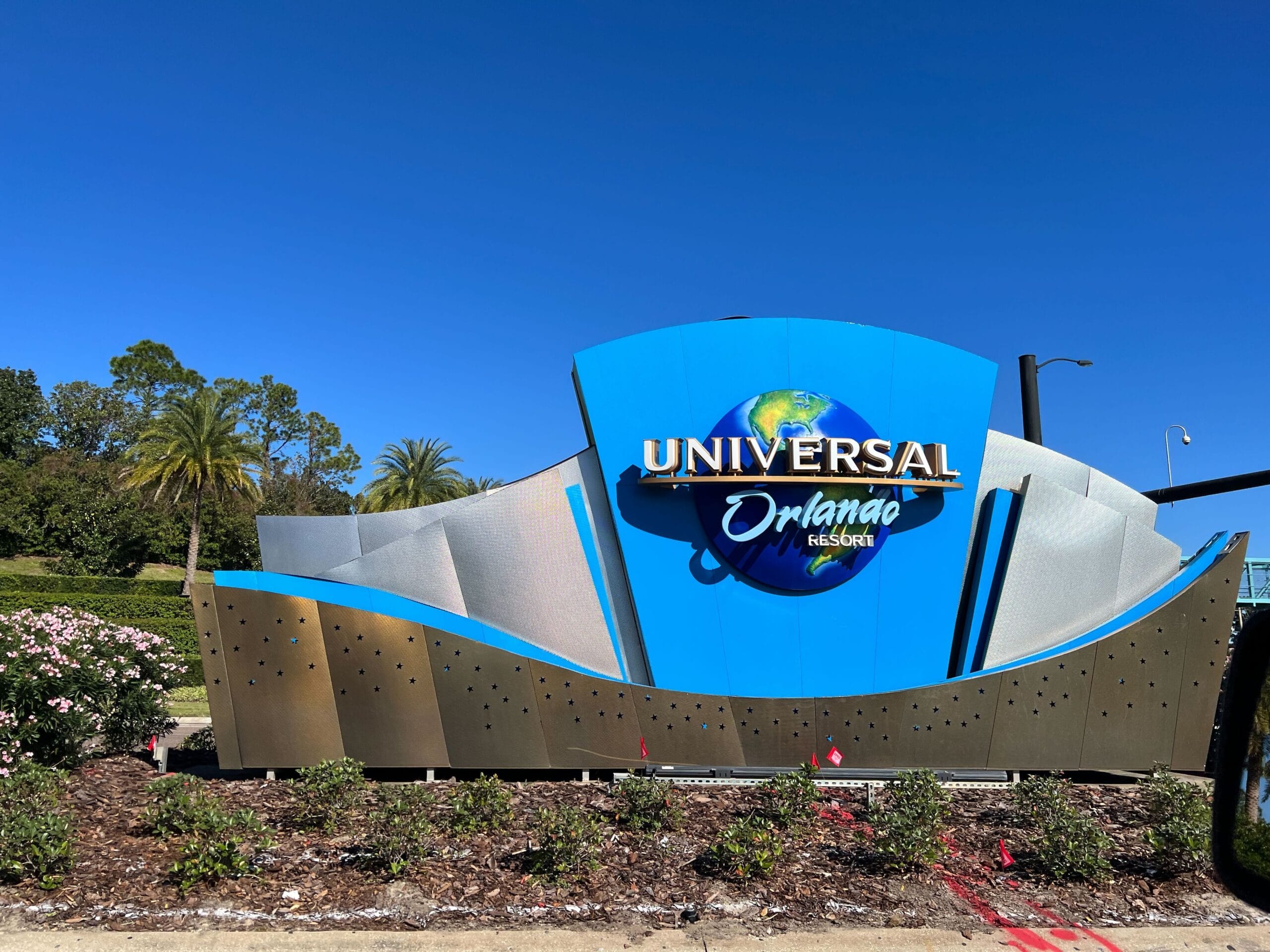 Universal Orlando Resort sign against a clear blue sky surrounded by greenery and flowers.