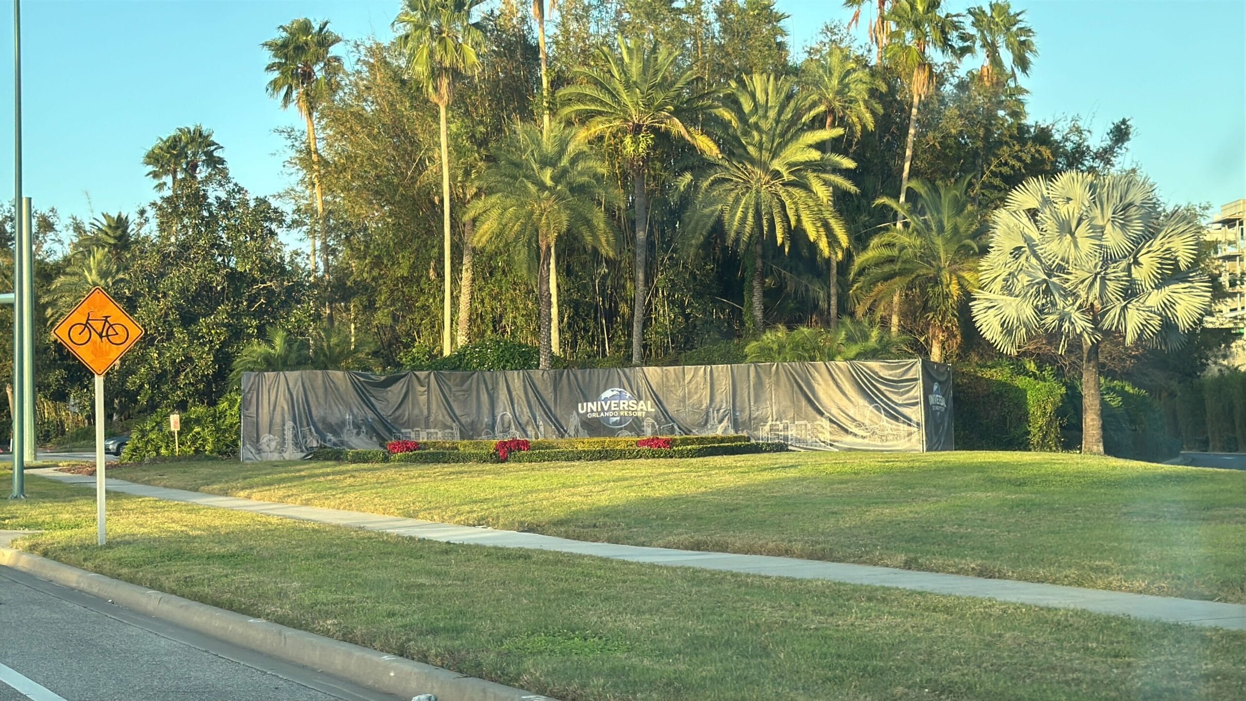 A street view with a universal sign covered with a tarp surrounded by palm trees and a bicycle crossing sign on the left.