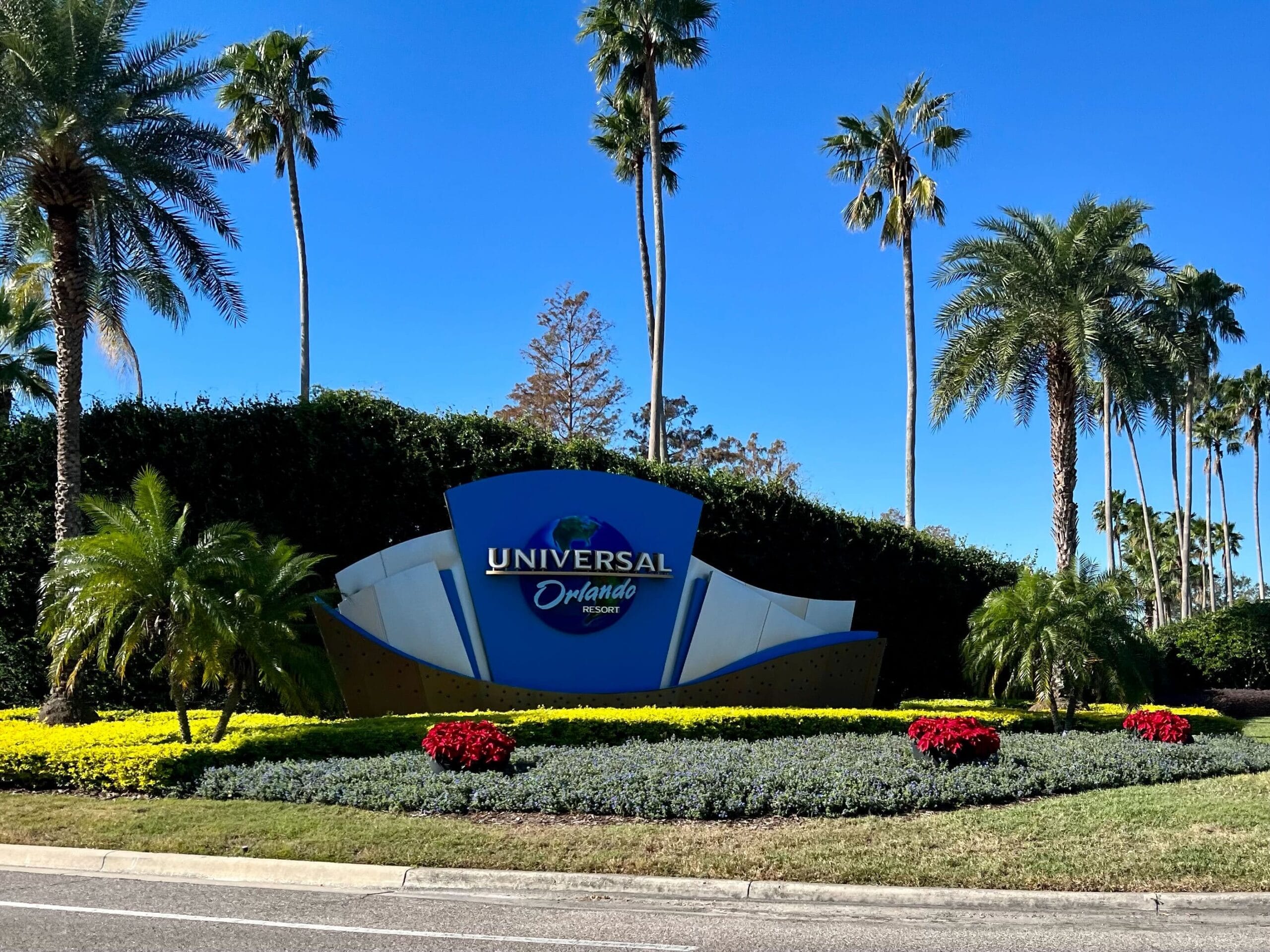 Entrance sign for Universal Orlando Resort surrounded by palm trees, bushes and colorful flowers under a clear blue sky.