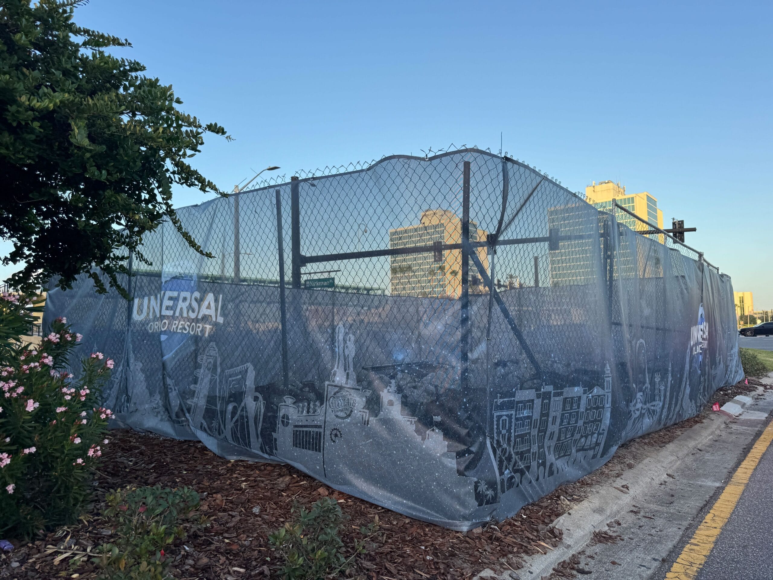 Chain link fence with universal branding and graphic images, next to plants and a road in an urban setting.