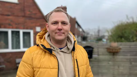 Karolis Arlauskas can be seen as he is happy outside of his property. He wears a hoodie with a yellow coat. His red fence and fence can see the background.