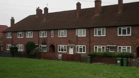 A number of red bricks terraced houses with suitable roofs and several chimneys. Each house has white windows, small front gardens surrounded by wooden fences, and green wheelie waste buckets. The foreground has a grass area.