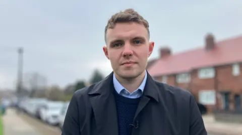 Charlie Davis can be seen forward. He stands outside in a residential street with a blue jumper and a shit with a marine coat. Rotbricked houses can be seen in the background and a number of parked cars.