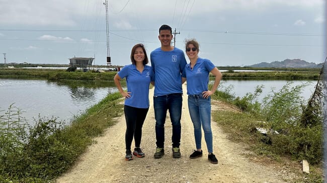 Three people stand together next to shrimp ponds.