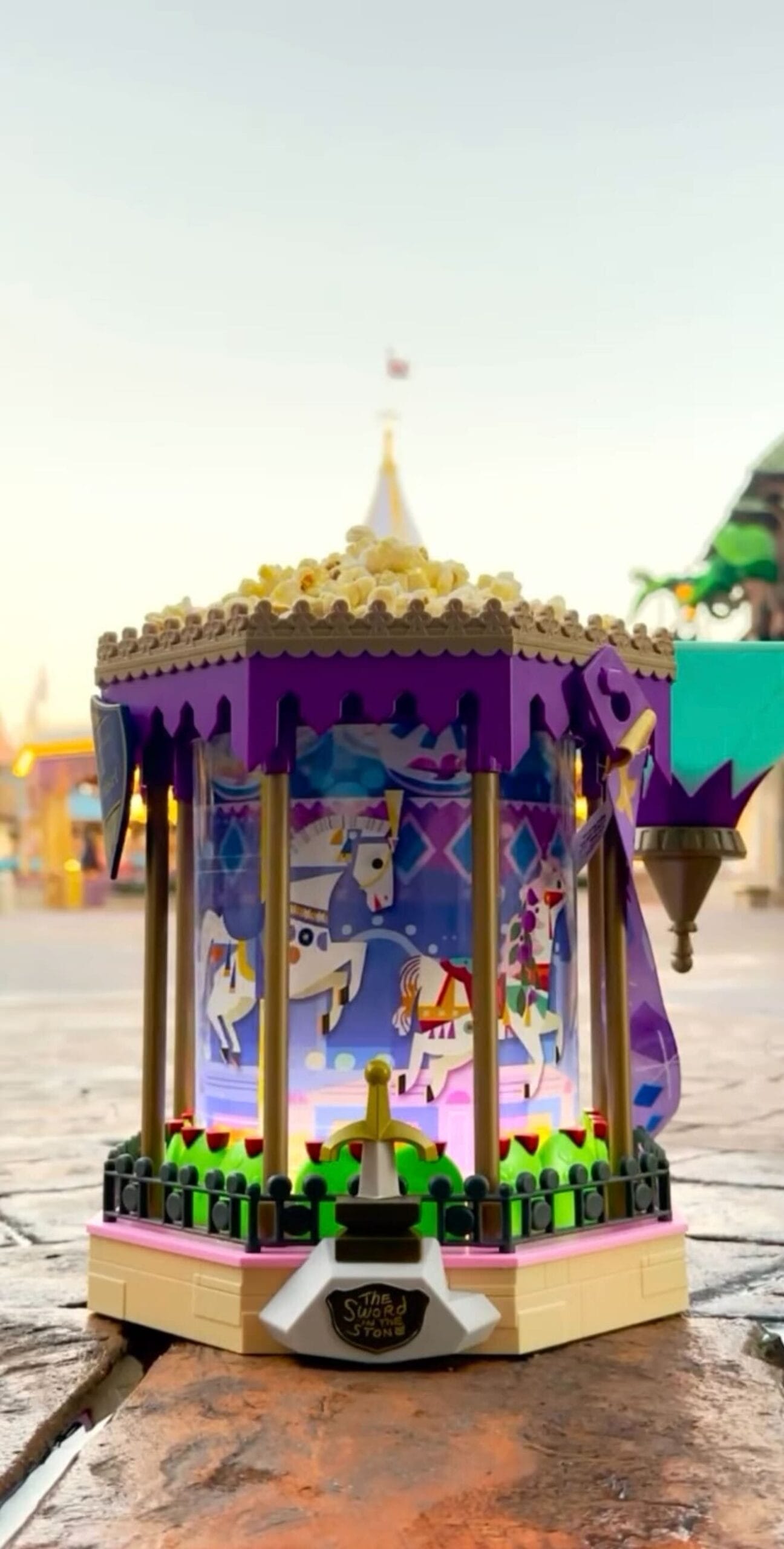 A small, decorative carousel with purple cut and horse detonator is placed on a brick surface. The background is an outdoor environment with a clear sky.