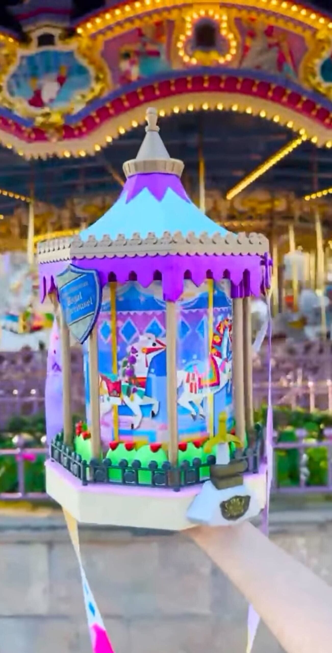 A hand contains a colorful carousel -shaped object with complicated details. A real carousel is blurred in the background.