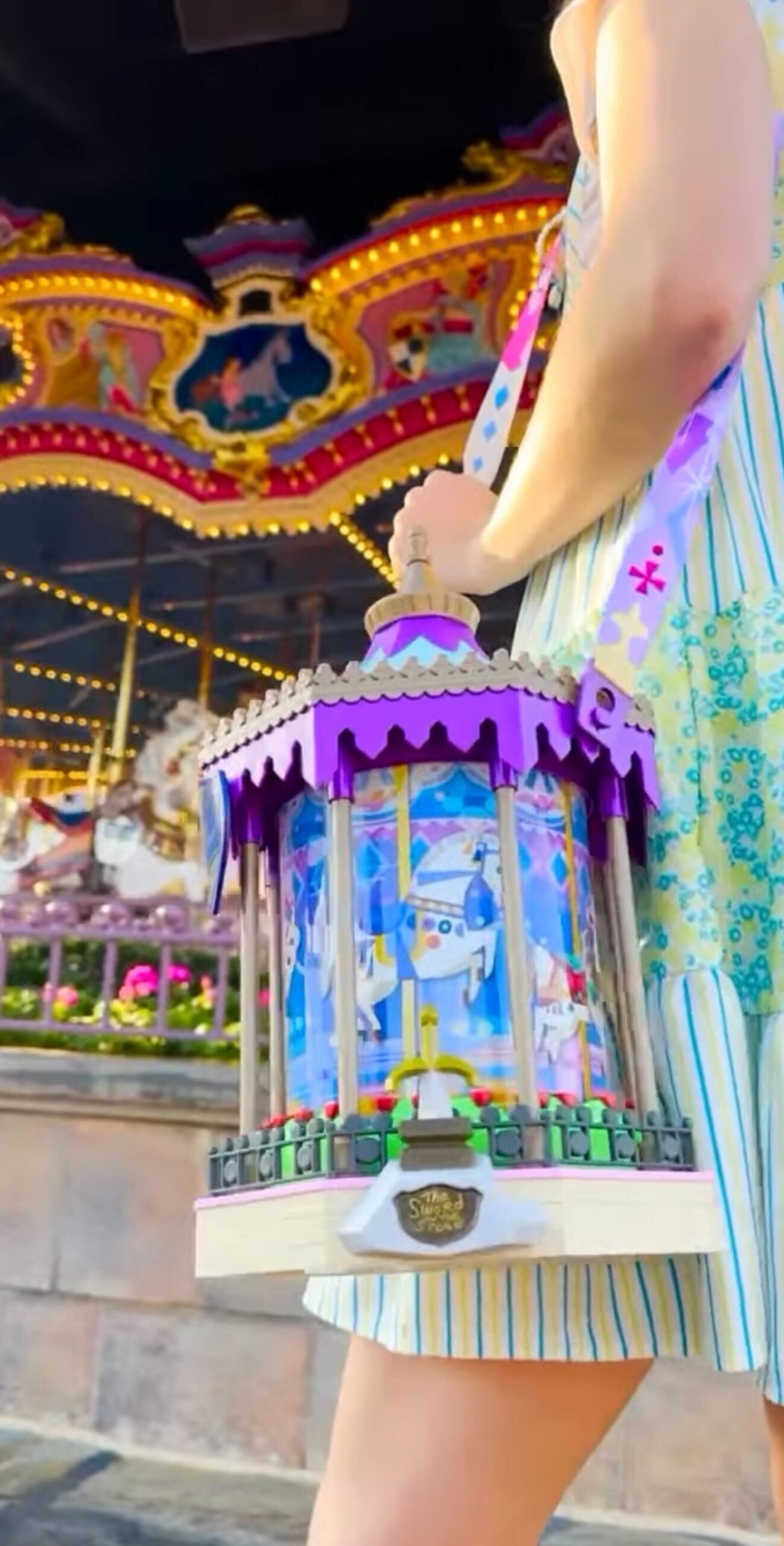 Person who holds a decorative bag that is shaped like a carousel in front of a real carousel.