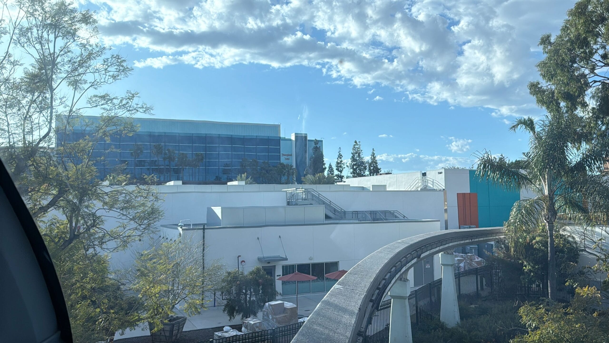 The Disneyland Monorail route with support pillars is gracefully through a scenic area full of trees, in addition to modern buildings under a clear, partially cloudy sky. Recently reopened, it offers a seamless journey through scenic and urban landscapes alike.