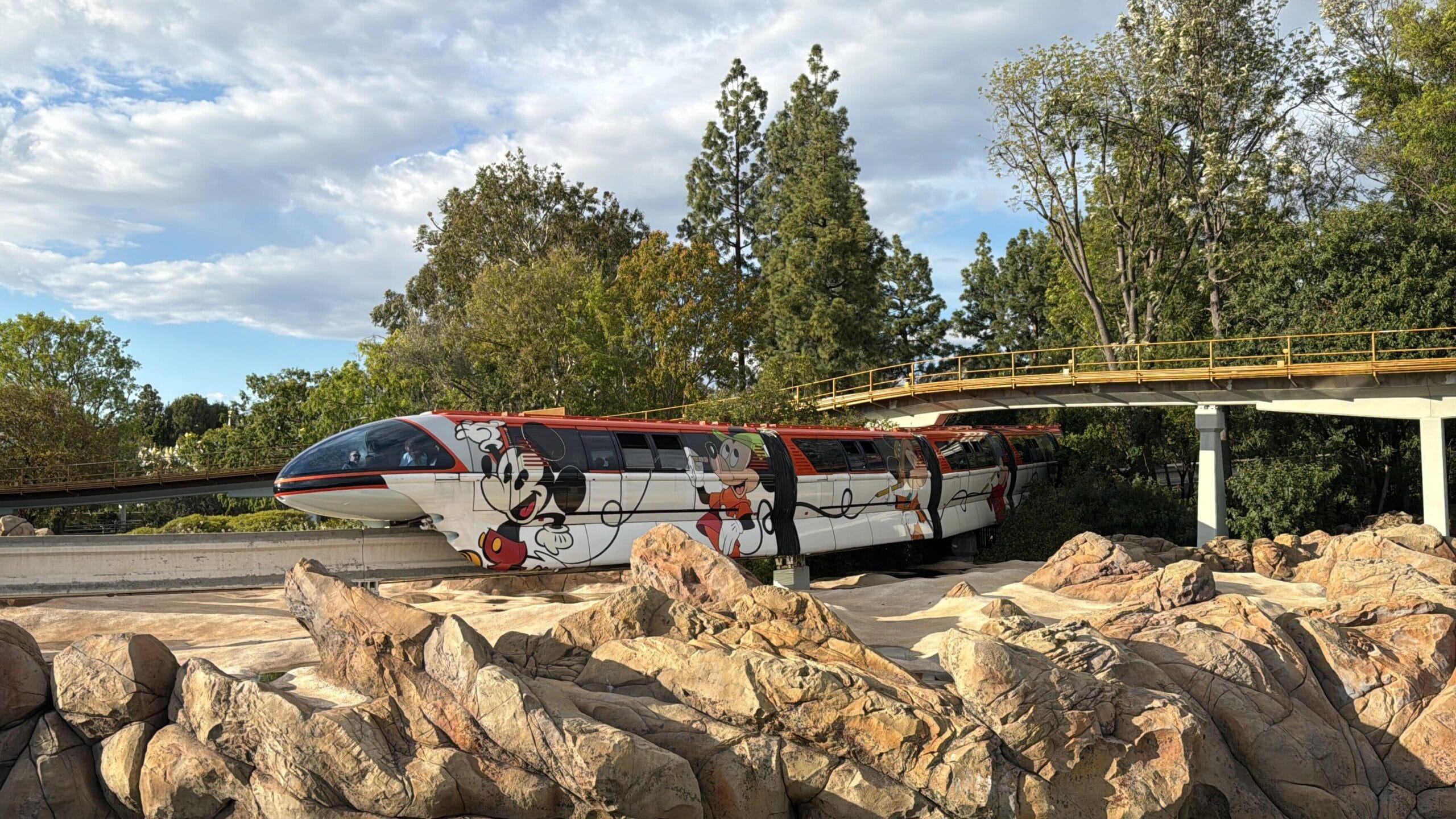 The Disneyland Monorail, decorated with colorful cartoon character designs, glides along its reopened route, surrounded by lush trees and rocks under a partially cloudy sky.