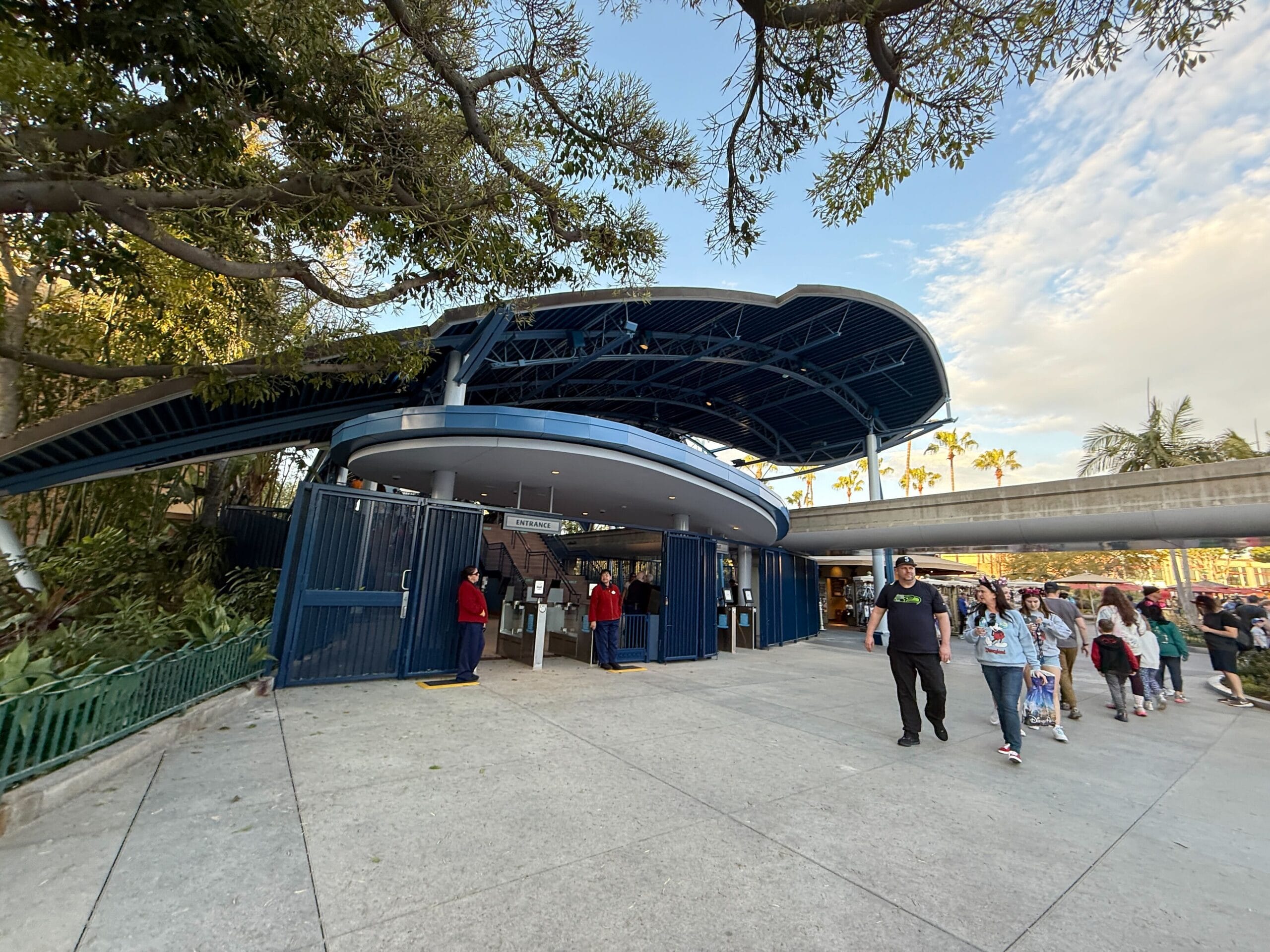 The people walk near a large blue -white curved structure, which is reminiscent of the Disneyland Monorail with its slim design and entrance gates, all of which are framed by trees and a clear sky.