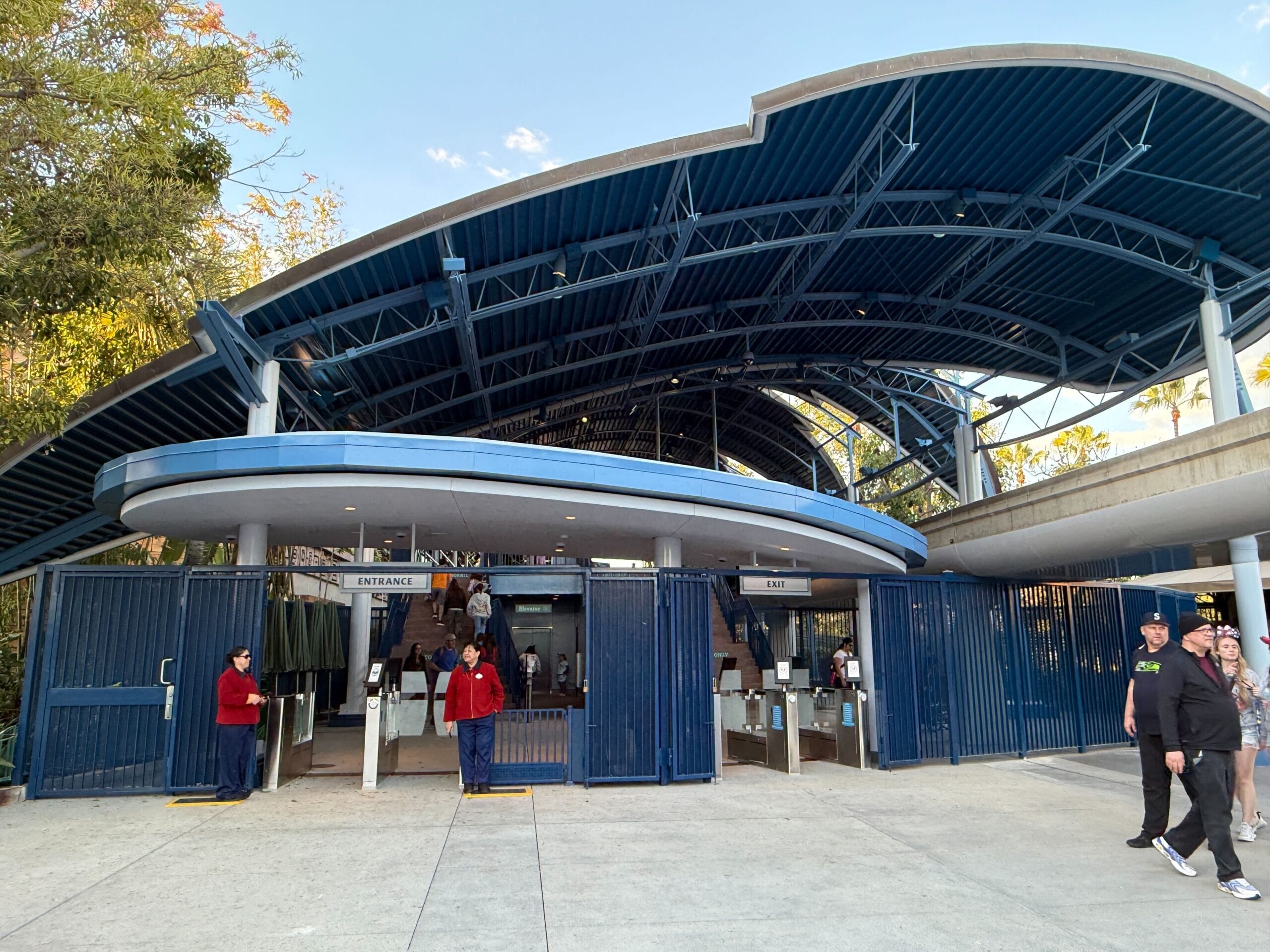 The outdoor entrance gate with a slim blue canopy and metal hinge welcomes visitors while waiting near the legendary monorail of Disneyland. Lush trees offer a picturesque backdrop.