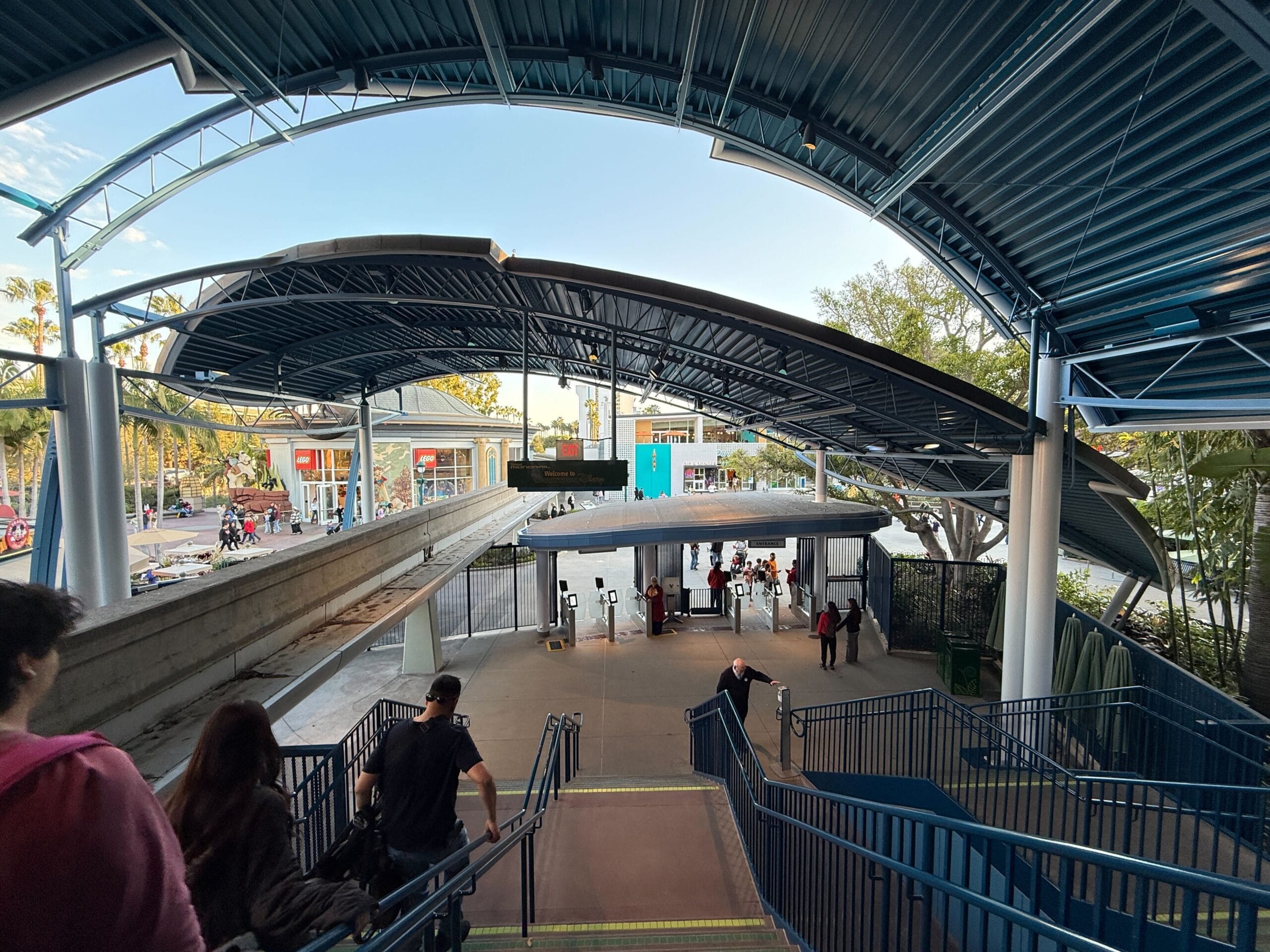 People who descend the stairs to a bus railway with a covered waiting area; Trees and a street with cars are visible in the background and are reminiscent of the ordinary river at the entrance to the Disneyland Monorail.