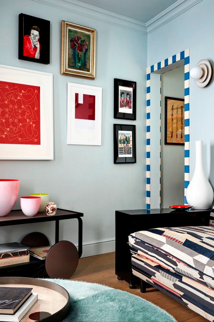 A room with light blue walls has a gallery with a framed work of art, a striped door, a patterned bench, a black table with decorative vases and a round carpet