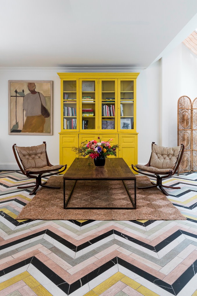 A large studio room with a tiled floor. The tiles that are created in a strong Chevron pattern are white, pink, yellow and gray. At the other end of the room there is a coffee table and two chairs with a bright yellow bookshelf.