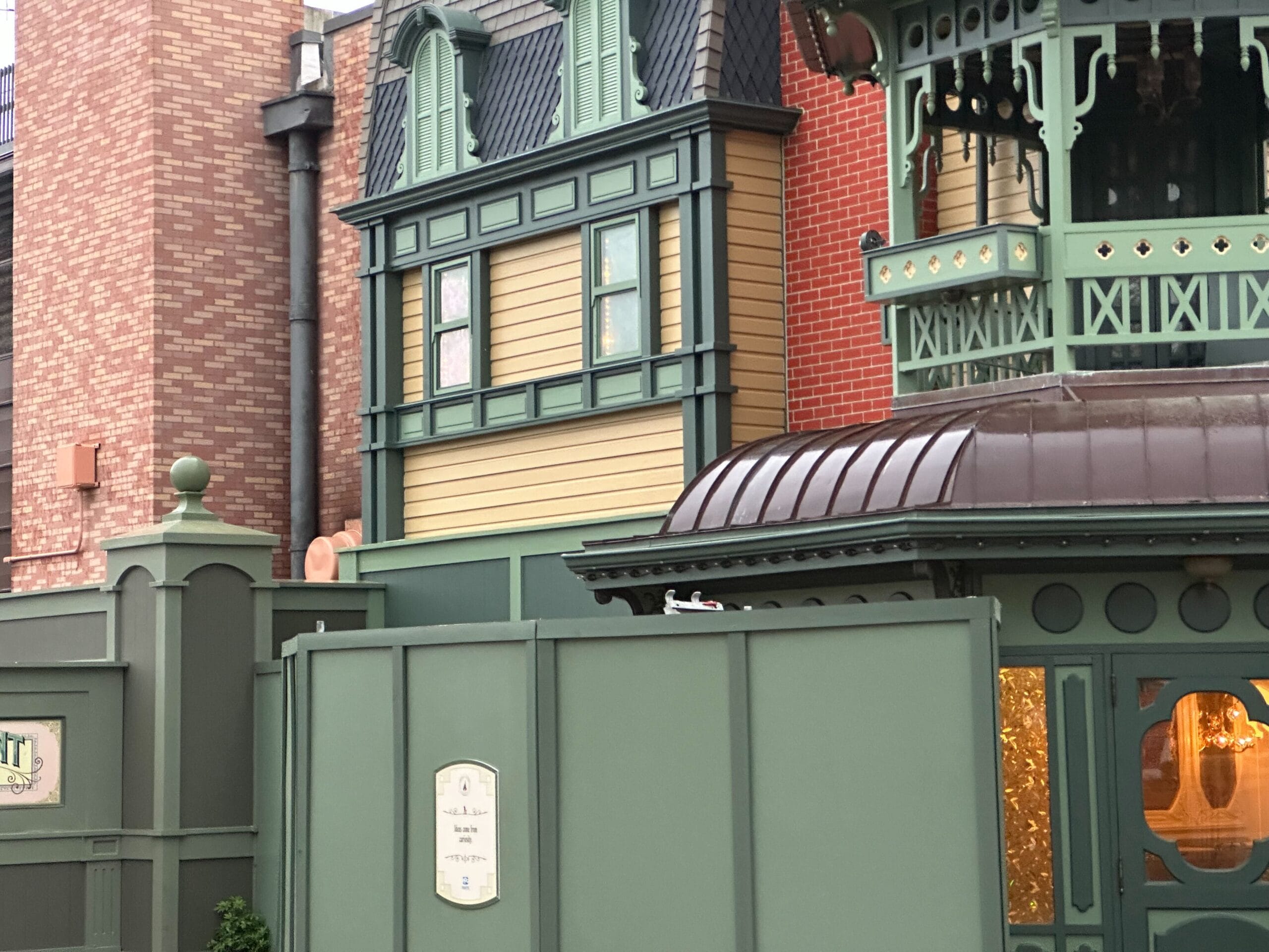 A construction site with a green fence in front of a building with a green expression and yellow siding. A partially visible wall is behind the building.