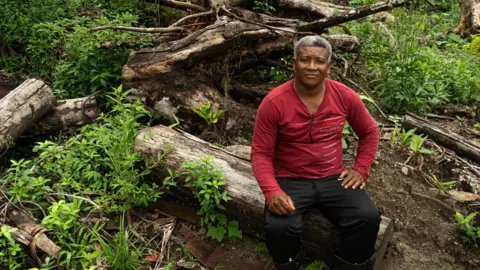 BBC / Paulo Koba Claudio Verequete sits on a felled tree with a red jumper. He has short gray hair and looks into the camera