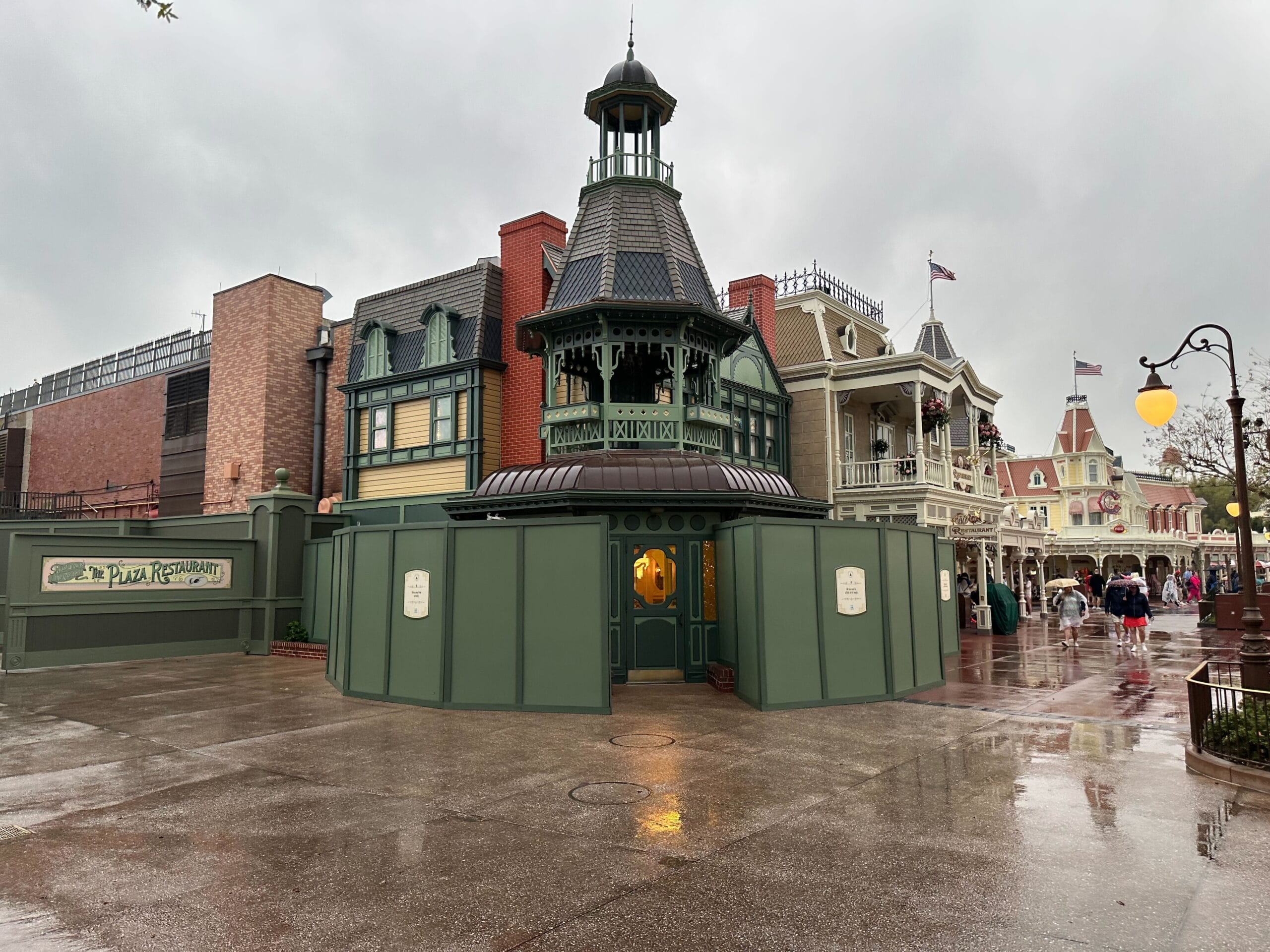 Rainy day in a theme park with buildings in the Victorian style and construction barriers "Plaza restaurant." Few people go on the wet sidewalk.