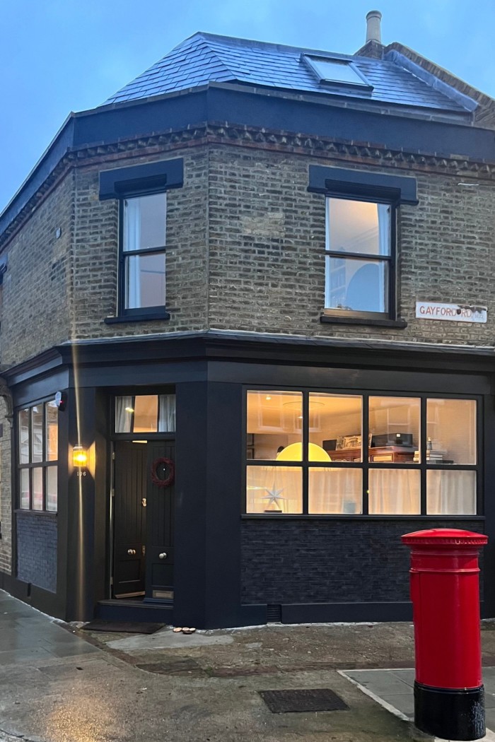 A cornerstone building with black window frames and doors is illuminated from the inside, with a red postciste on the sidewalk and a street sign by reading “Gayford RD W12