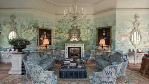 A large living room has hand -painted landscape paintings, decorated blue furniture, a chandelier, a fireplace with a mirror and portraits that are framed in gold