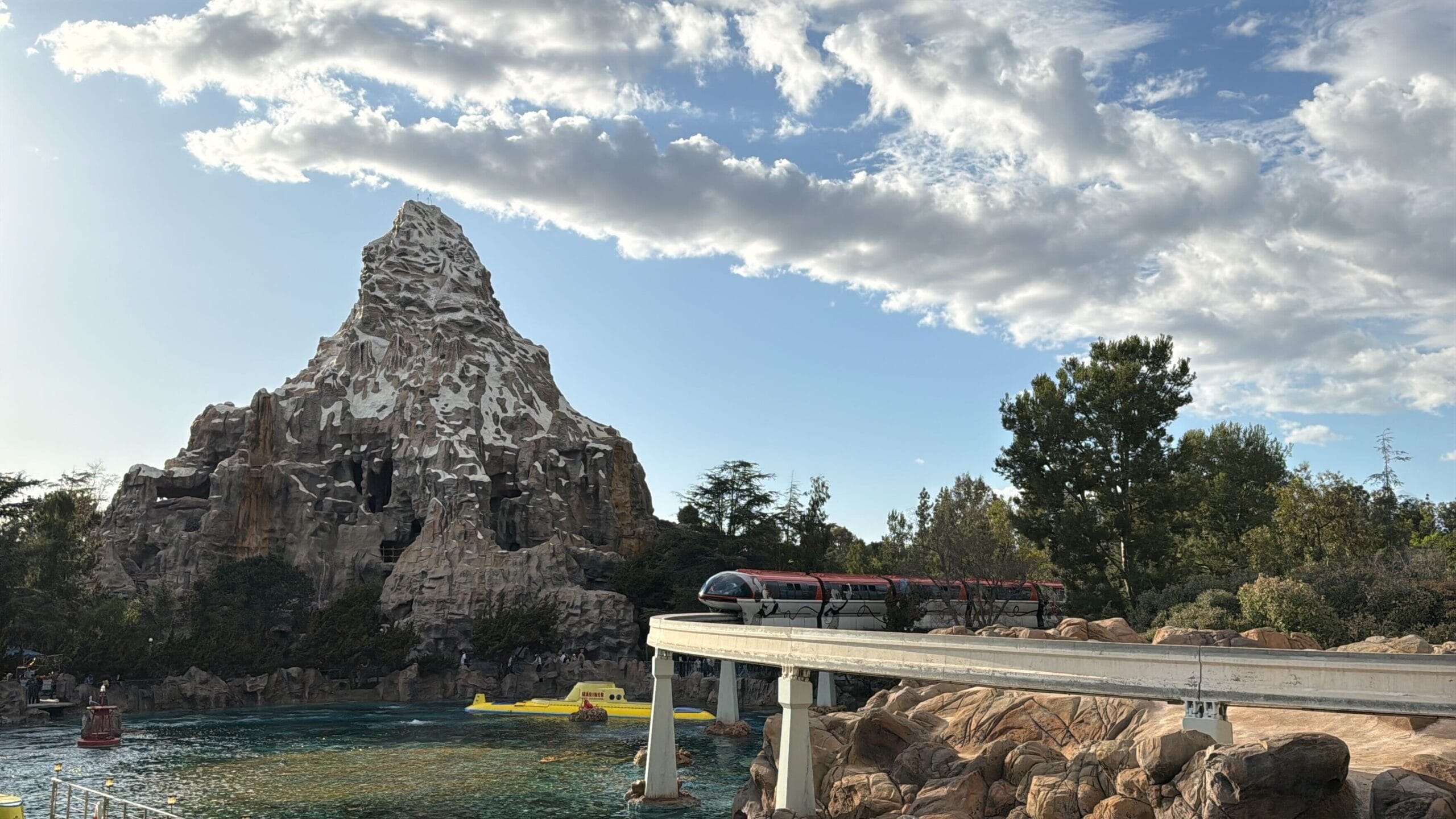 A Red Disneyland Monorail drives trees on a raised route in front of a mountain while a U boat swims in the water down. The sky is partially cloudy and captures the magic because it welcomes visitors again.