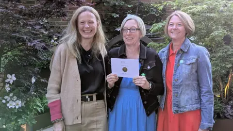 Jen Donnelly smiles three women. A woman in the middle with glasses holds a paper envelope with the medal.