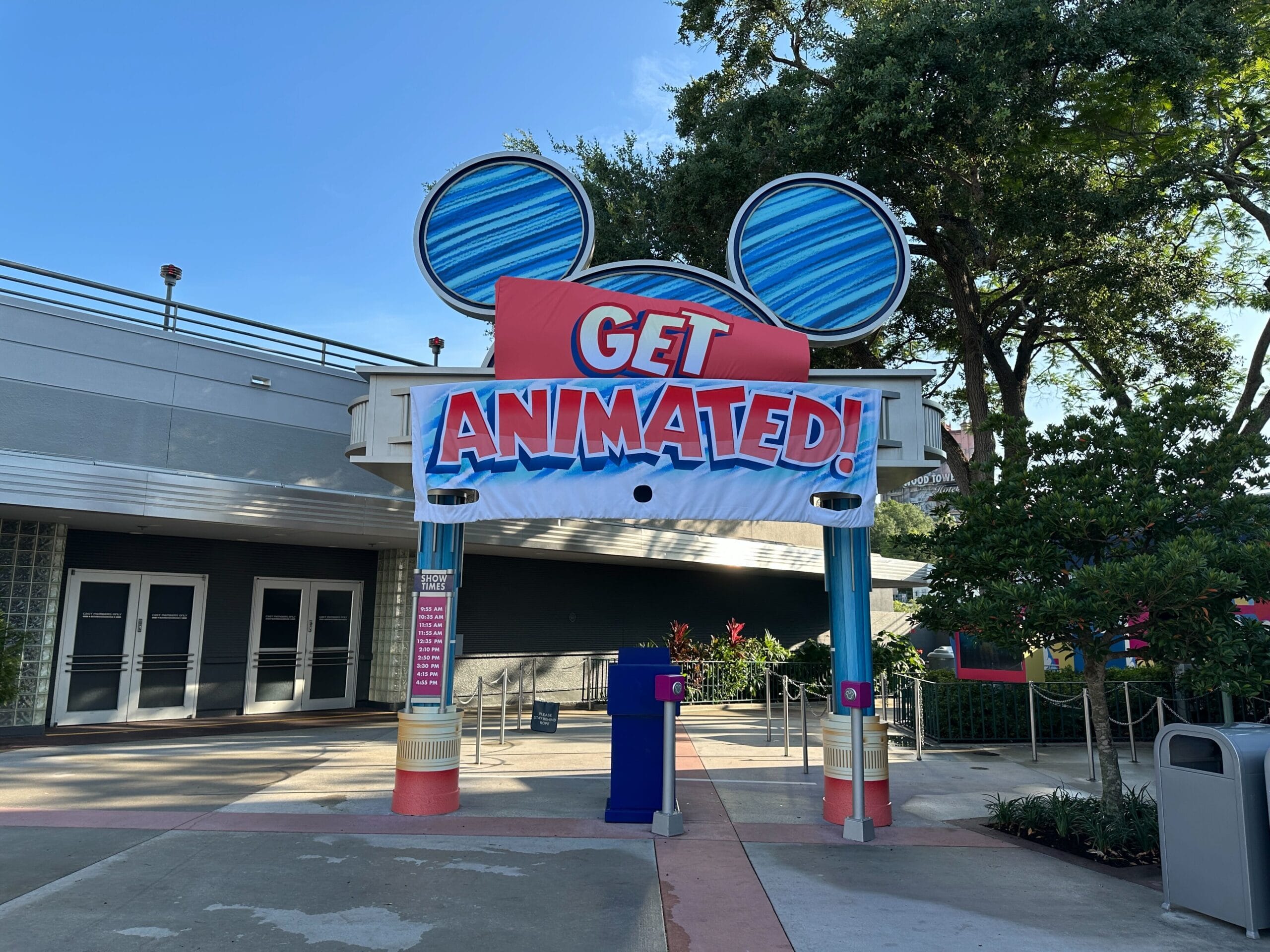 Colorful "Get animated!" Sign with Mickey mouse ears over an entrance in a theme park, surrounded by trees.