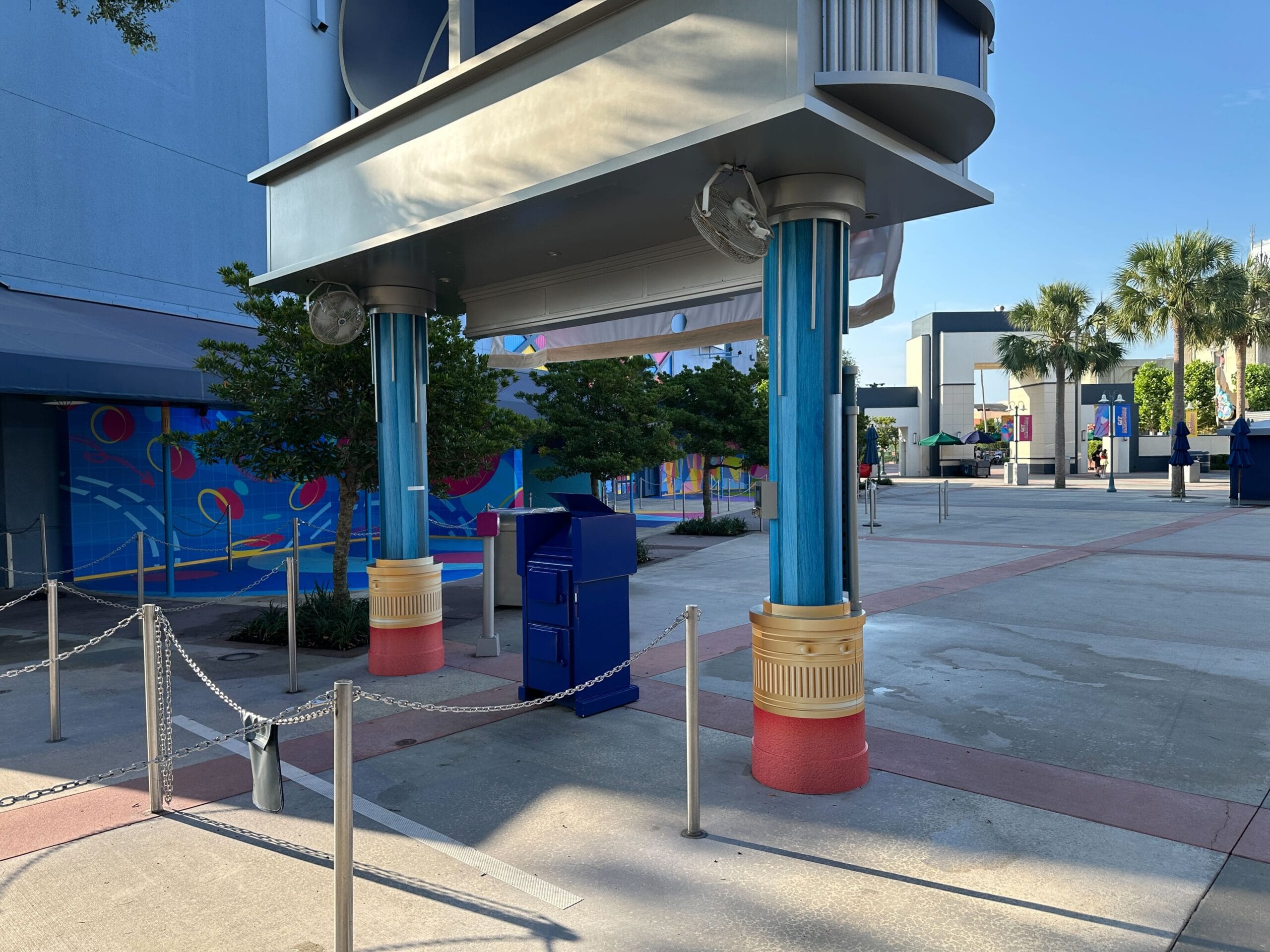 Colorful outdoor area with blue columns, trees, a chain barrier and a painted wall painting in the background.