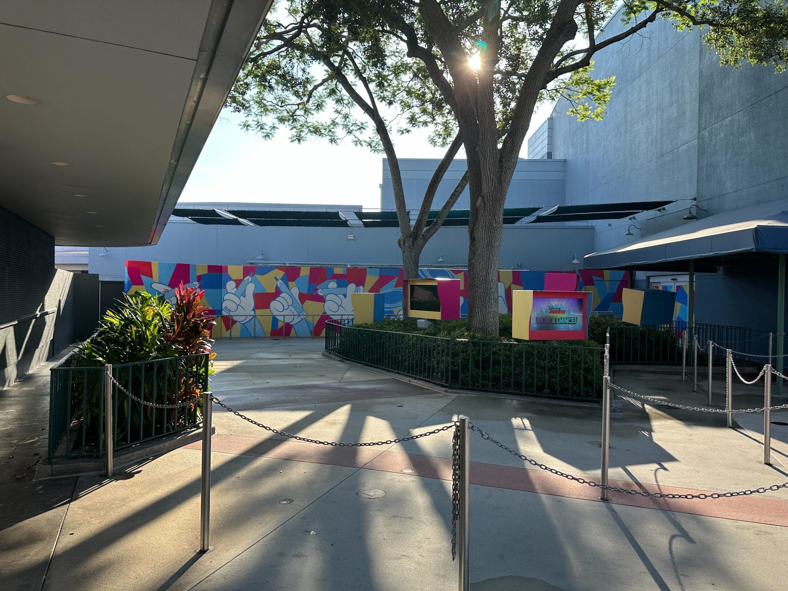 Colorful construction wall with abstract art and elevated thread drives, surrounded by trees and cable barriers in sunlight.