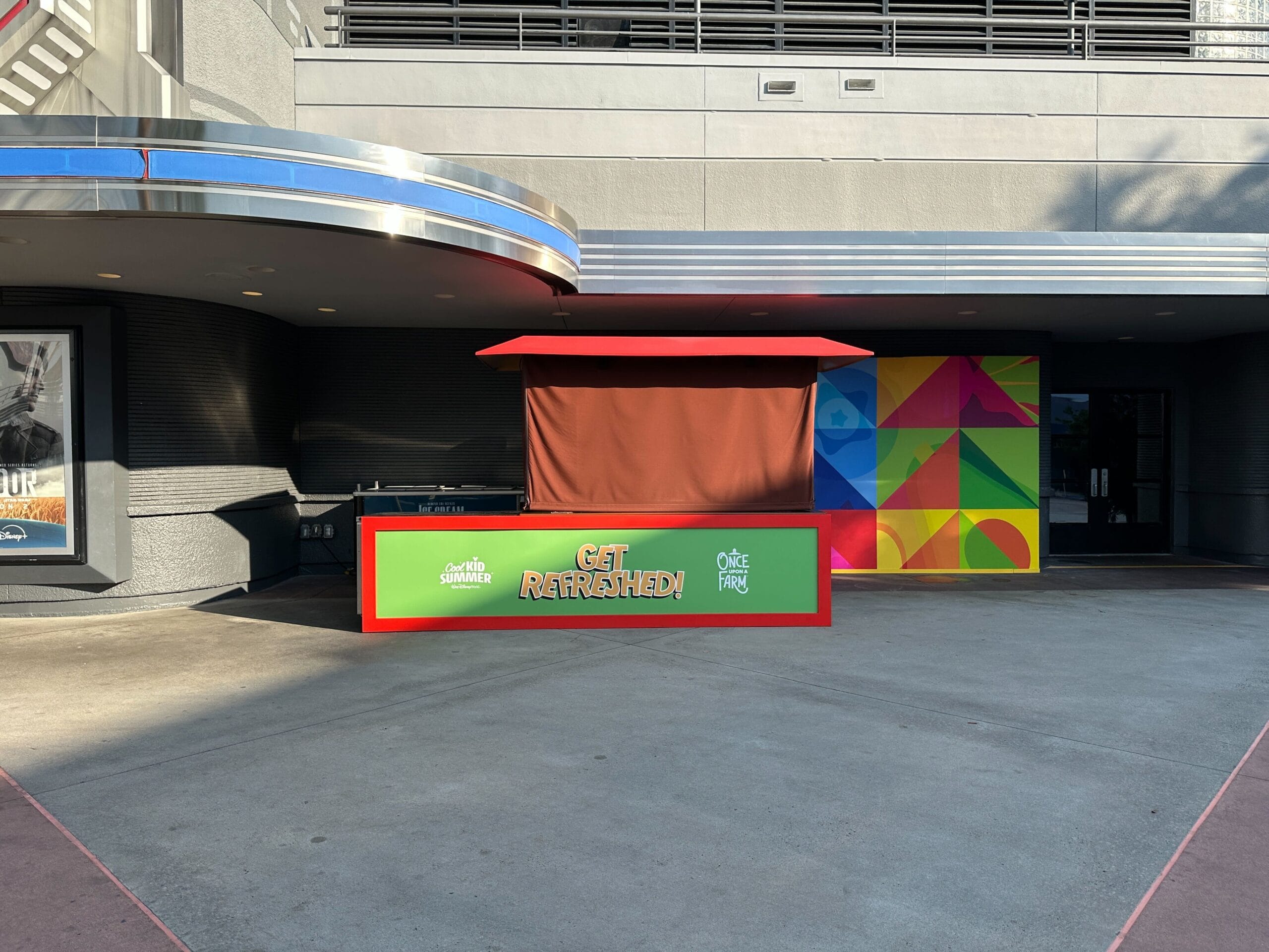A closed refreshment stand with one "Refreshed!" Sign under a building, with colorful panels on the right.
