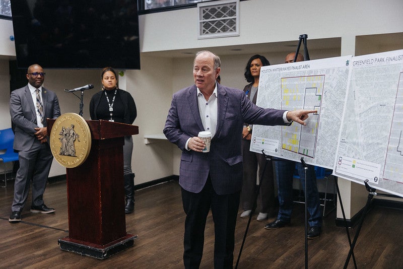 A white man in a gray suit that indicates details on a posterboard. There are four other people in the background.