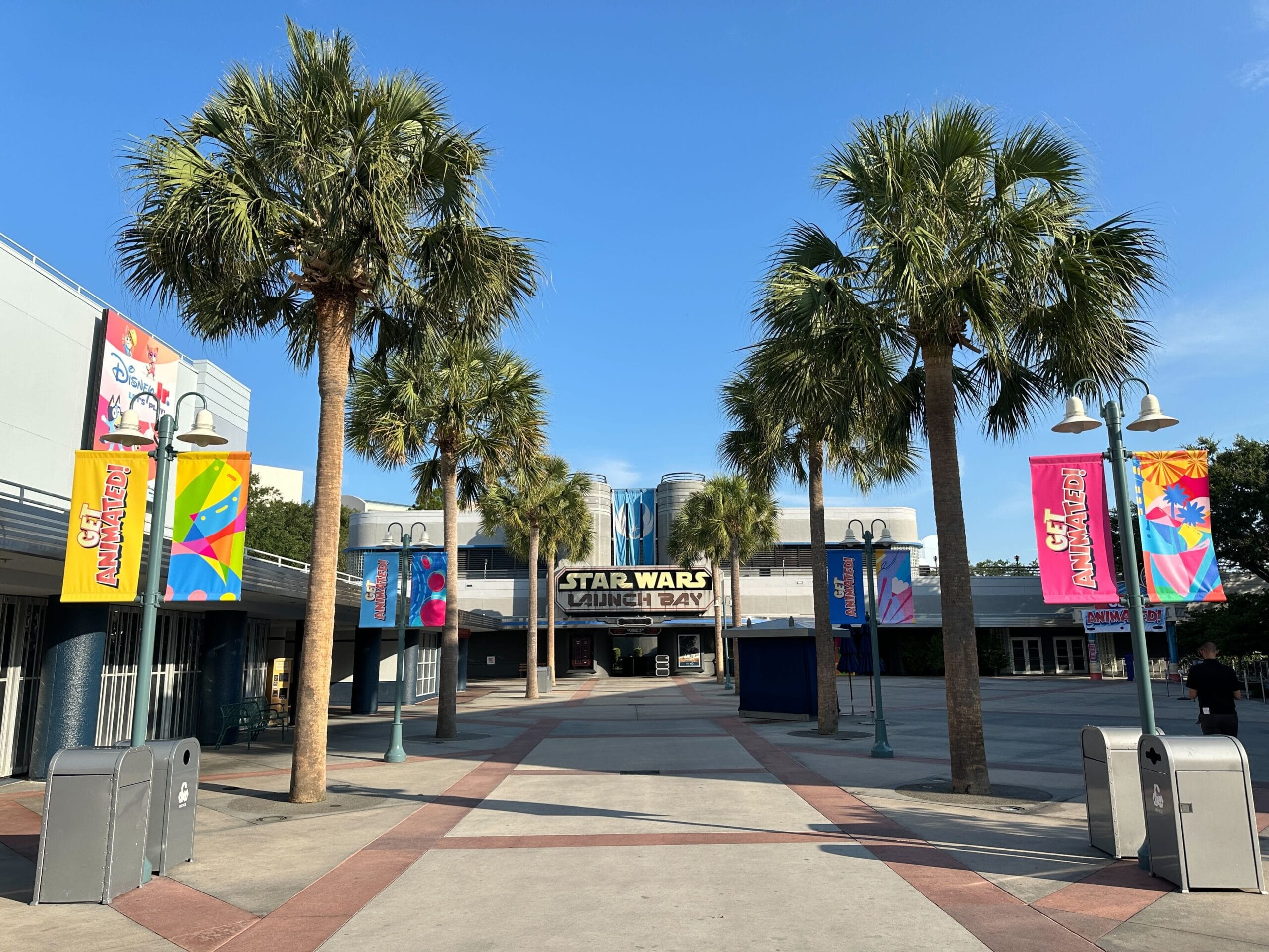 Palm trees line a sidewalk that leads to the Star Wars Launch Bay building under a clear blue sky.
