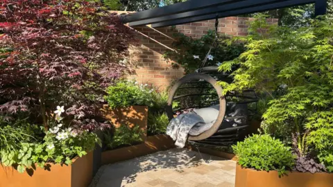 Jen Donnelly Different plant containers on different levels are full of green plants, some trees with purple leaves and white flowers. There is a hanging chair in the middle.