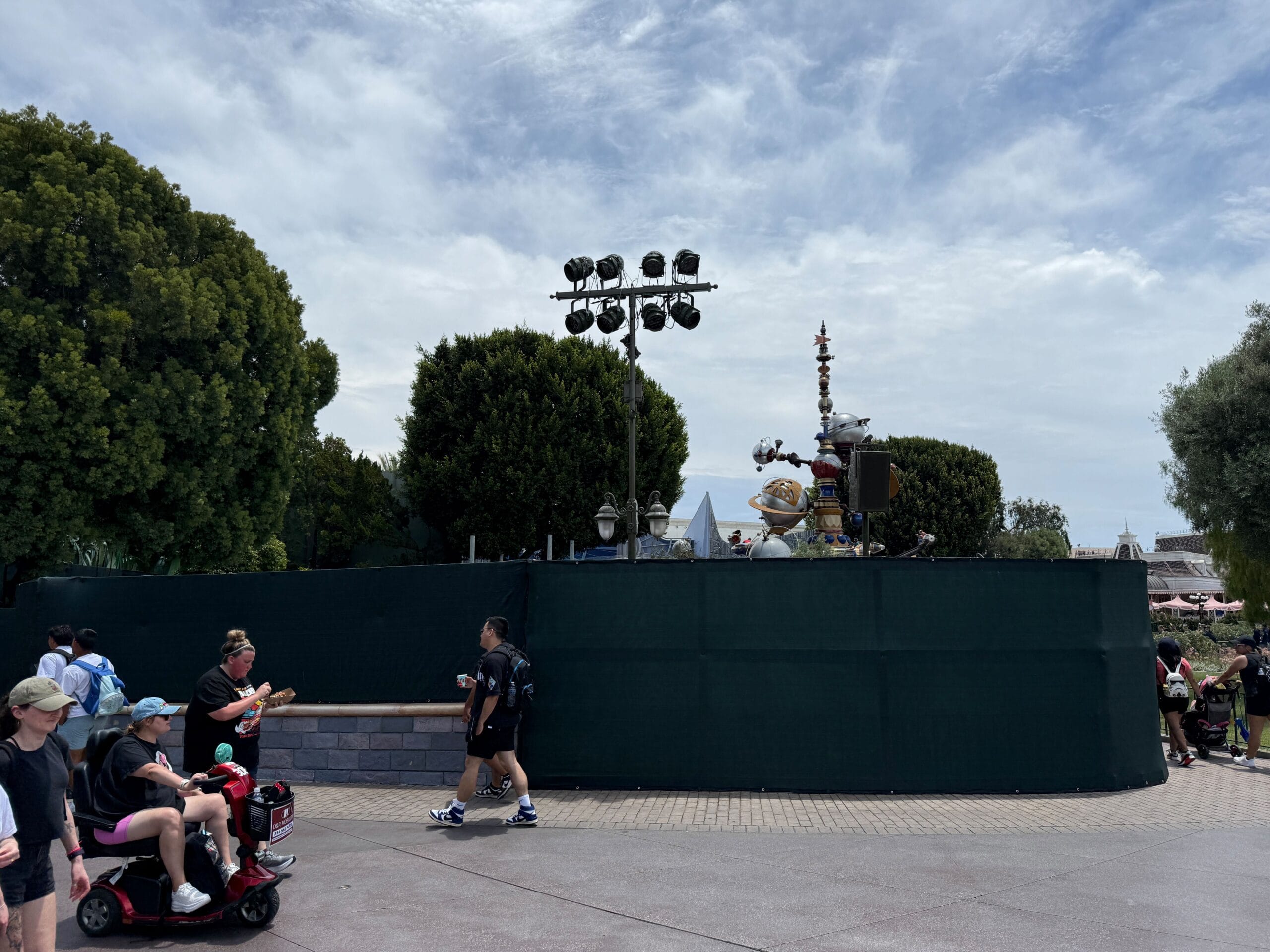 People walk past a green construction fence in a theme park, with trees and a spaceship visible in the background.