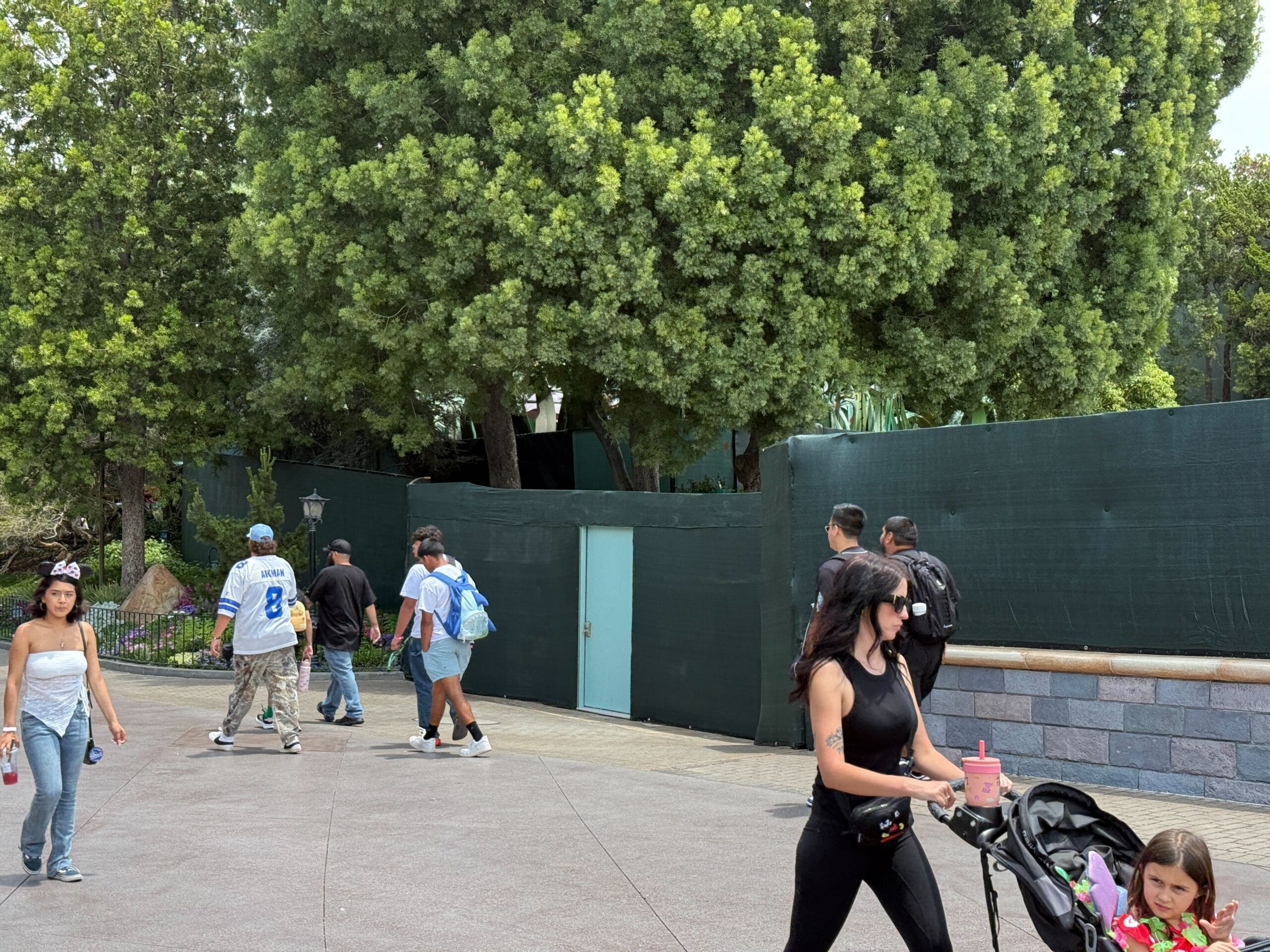 People walk past green walls and trees; A woman pushes a stroller and a girl goes next to her.