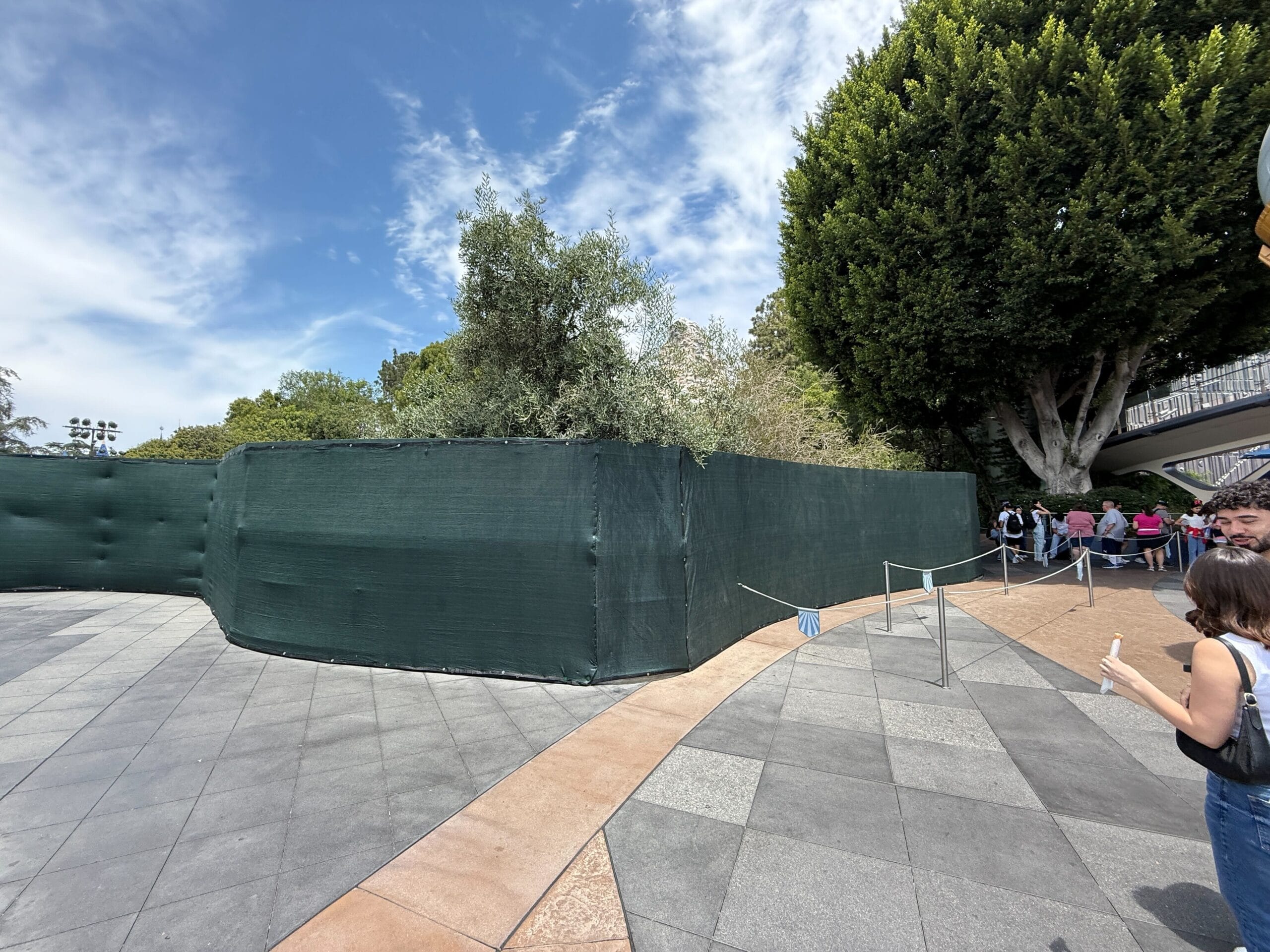 On a sunny day, green construction fences blocks an area near a series of people and large trees.