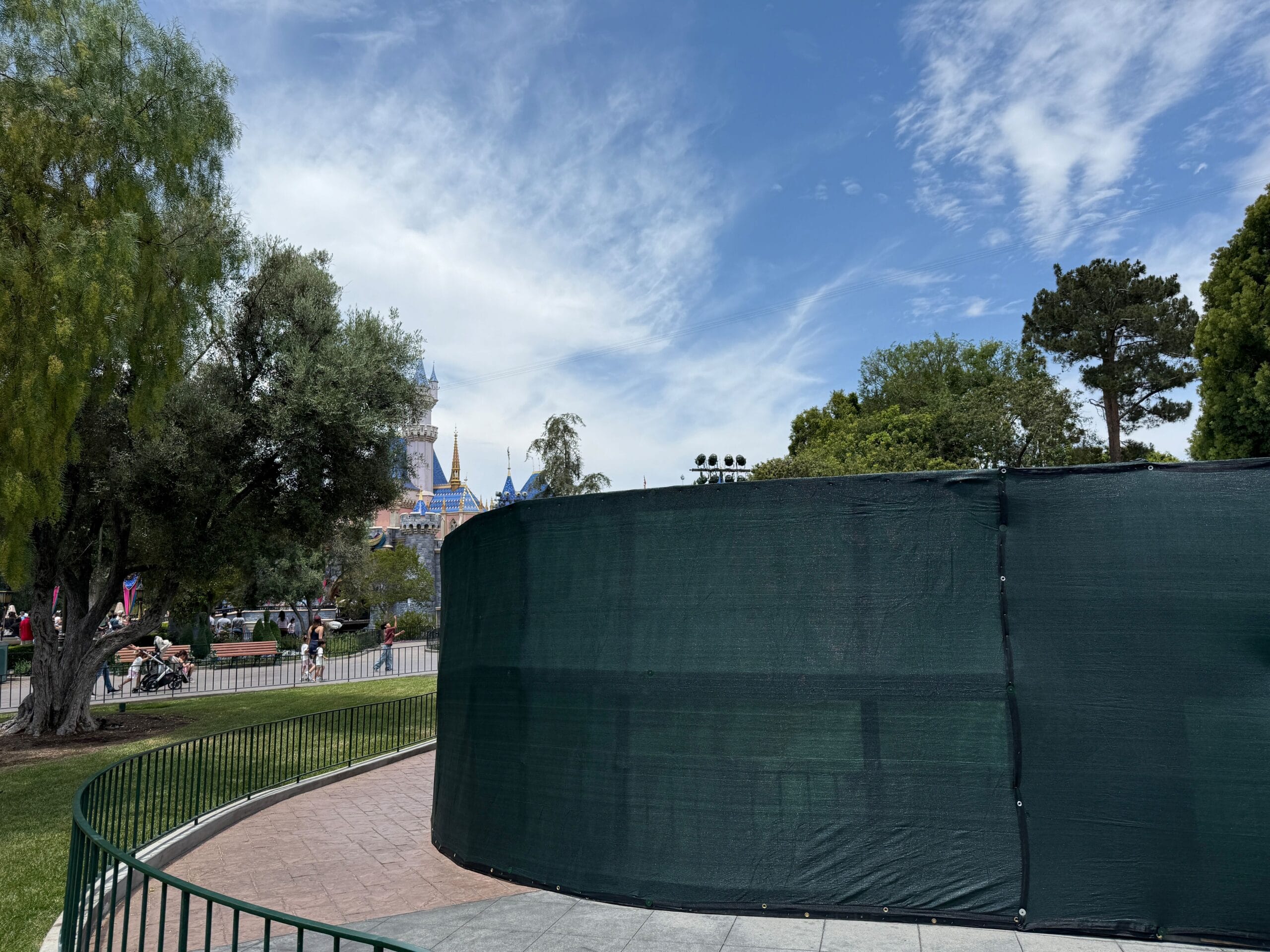A curved green construction fence blocks part of a sidewalk in a park; A lock is visible in the distance under a blue sky.