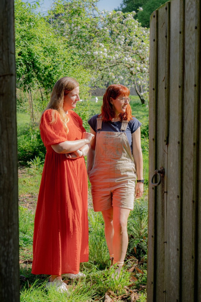 A woman in an orange dress and another who wears short dungarees is in a door with a view of the garden behind it.