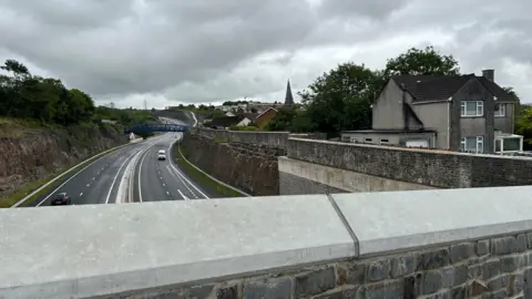 A view from a bridge on the heads of the valleys of the dual lane. On the right there is a large stone wall and John's house on a platform to the right of the wall. It is a cloudy day.