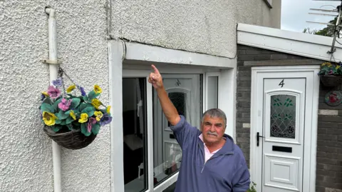 Daryl Wilkins, which points to a crack over his head in the outer white render of his house. He wears a blue sweatshirt and has gray hair and a gray mustache. He stands in front of a window with his front door to the right and a hanging basket on the left.
