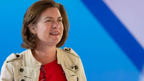 Getty Images Eluned Morgan looks into the distance. She wears a red top and a military style military jacket. There is a blurry blue background behind her.