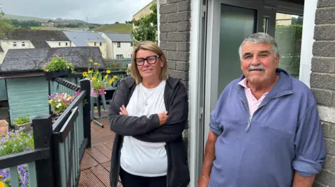 Julie and Daryl Wilkins stand in front of Daryl's house, Julie with crossed arm. He leans against the wall that is open next to him. Julie has blond hair and wears glasses, a white t-shirt and a black jacket. Behind them can be seen with flower planters, with a series of terrace houses in the background.
