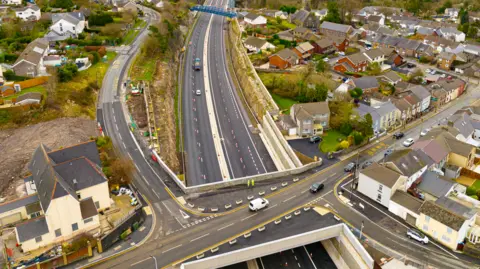 FCC building an air view of the heads of the Road Road and John Watkeys' house. A bridge with a street on the side can be seen that is crossed over the double road underneath, with the cones down the middle of each road. On the upper level, a series of terrace houses can be seen on the right side of the road with a housing estate on the right.