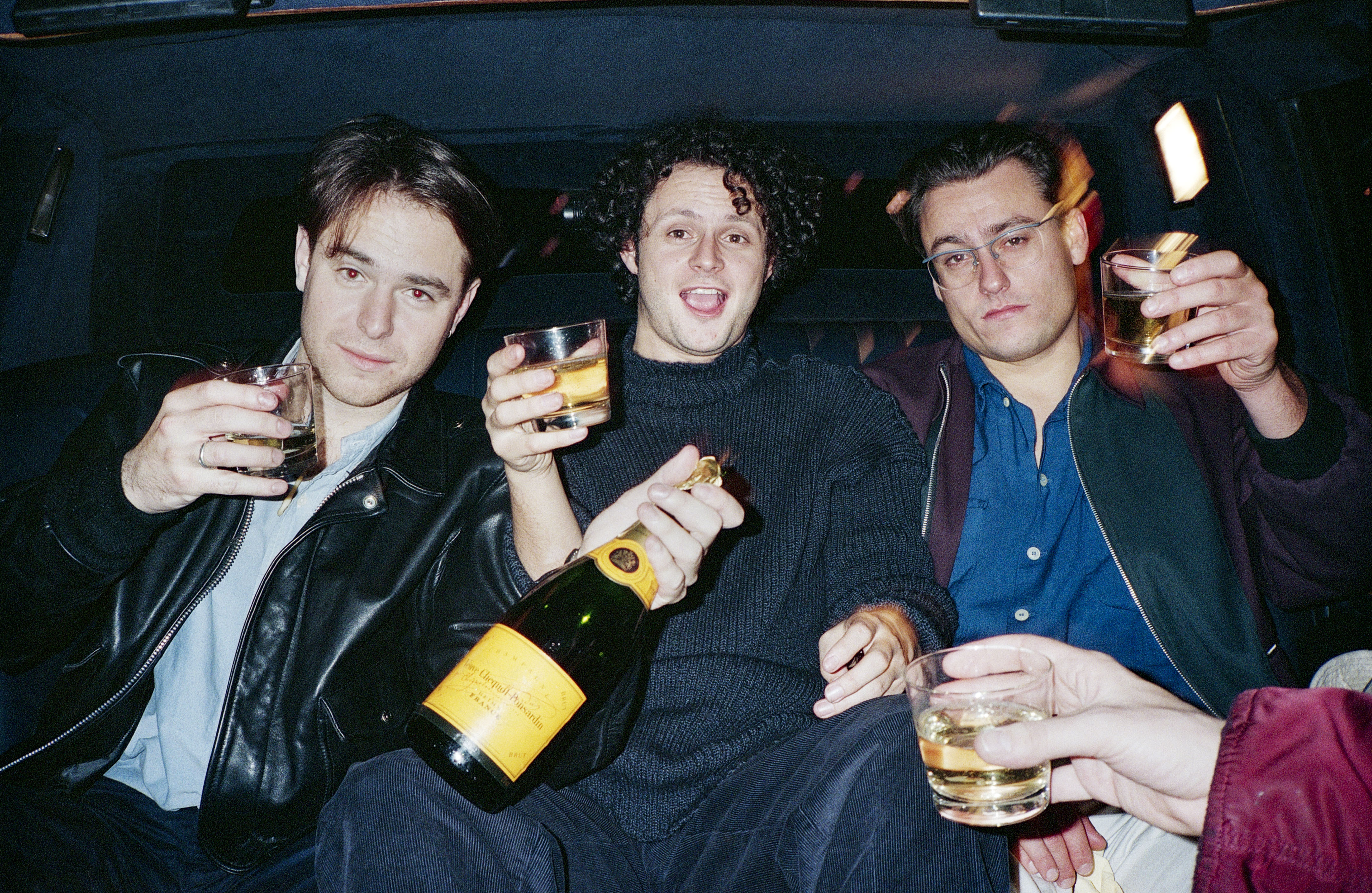 Three men celebrate with drinks in a car.