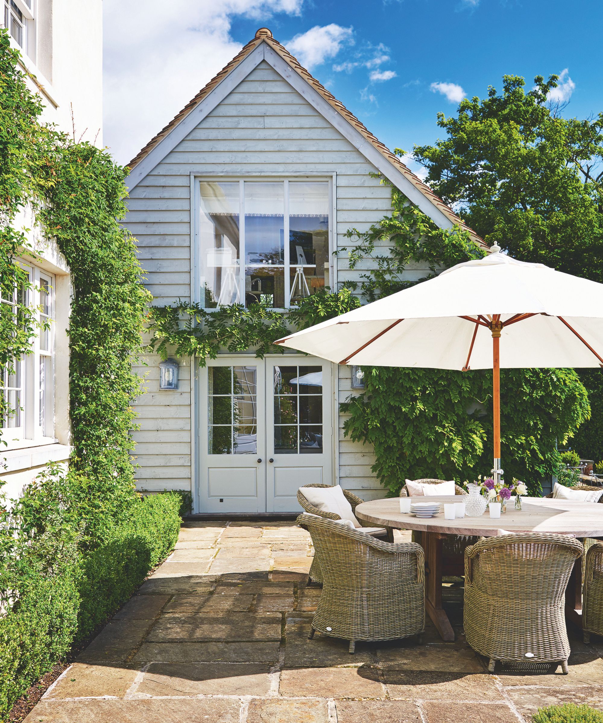 A sunny paved terrace with a white house, circular table with basket chairs and a large white, open umbrella