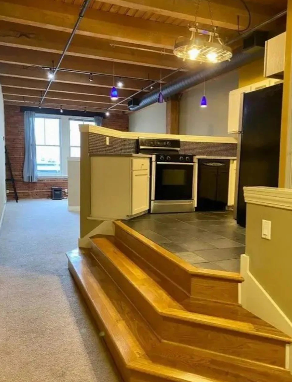 Kitchen slightly increased with wooden steps; has a stove, cupboards and overhead lighting in a loft-style room with visible rays