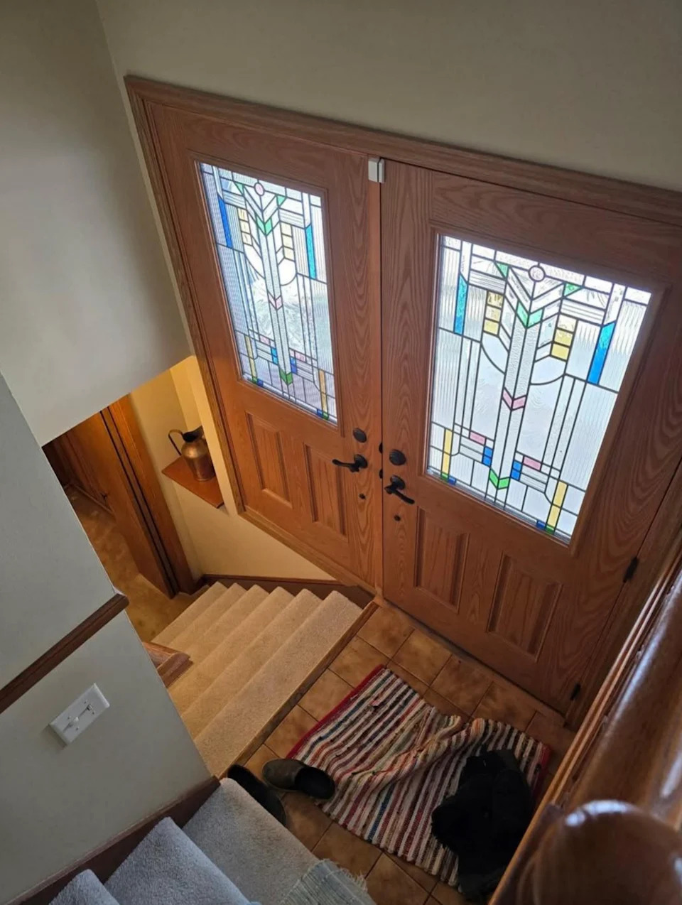 View of stairs of double wood doors with stained glass. A small carpet and shoes are at the entrance